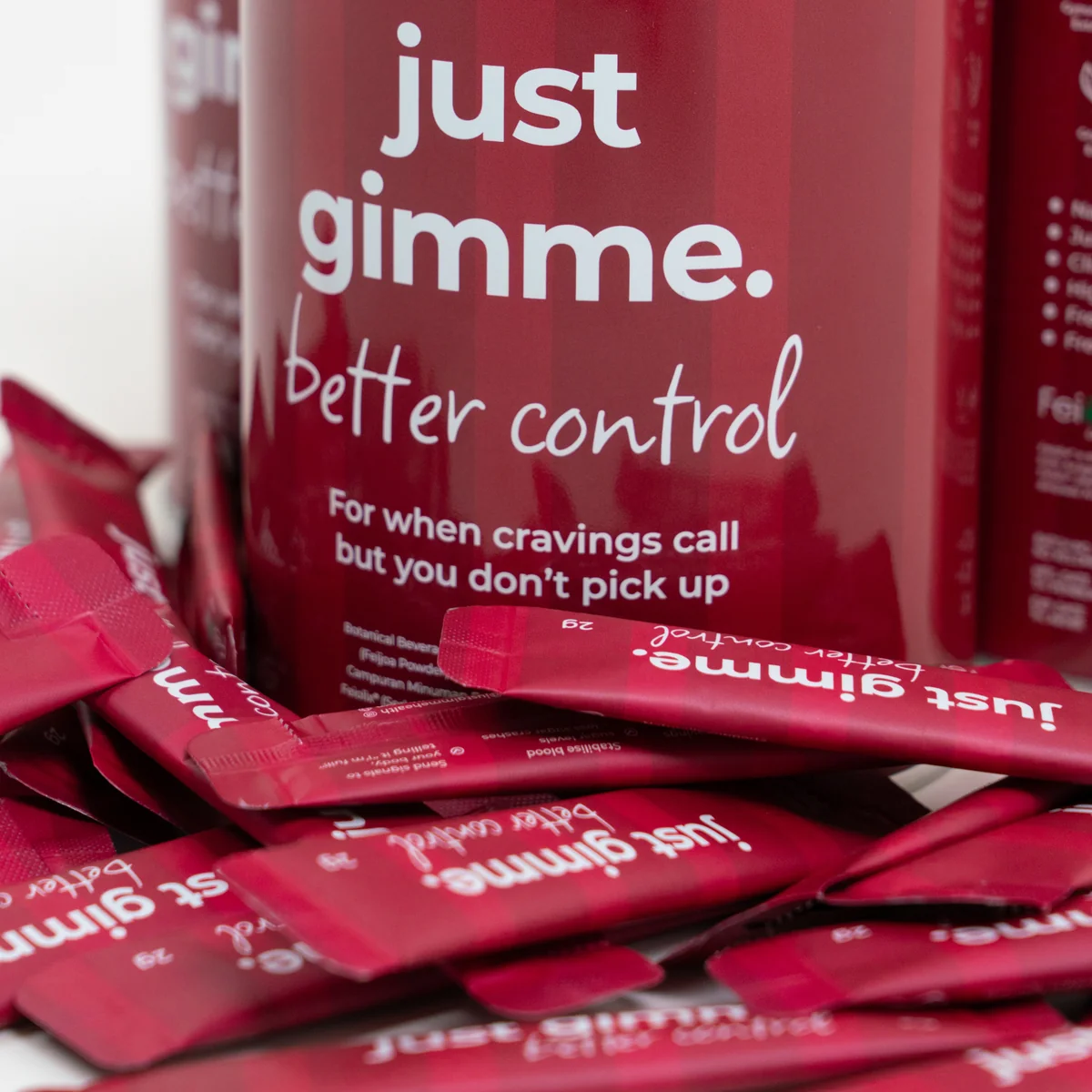 2048px_Web_PDP_11_Jan_2025_1 Red promotional bottle with white text reading 'just gimme' and handwritten 'better control,' surrounded by pink sachets on a surface.